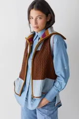 Chaleco de lana con cierre frontal completo y cuello alto. Presenta un diseño de bloques de color en tonos marrón chocolate y celeste, combinando secciones de tejido trenzado con secciones de tejido acanalado. Posee un corte asimétrico, siendo más largo en la parte posterior, y aberturas laterales en el ruedo.