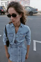 Vestido corto de denim azul claro, estilo camisa, con cuello solapa, escote pico, manga larga y cierre frontal de botones metálicos. Presenta bolsillos delanteros con tapa y bolsillos de plastrón en la espalda. La cintura está ajustada con elástico fruncido.