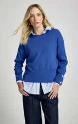 Sweater de punto color azul, de corte recto y ligero estilo holgado, con cuello redondo, mangas largas con puños ajustados por botones metálicos y dobladillo acanalado.