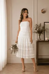 Vestido midi strapless color marfil, con diseño de paneles horizontales que combinan encaje y detalles de flecos. Presenta una falda con vuelo y movimiento.