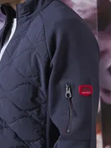 Campera azul marino con cierre frontal, cuello alto y bolsillos laterales con cierre. Tiene un parche rojo con cierre en la manga izquierda y un diseño acolchado en la parte delantera.