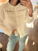 Blusa de manga larga color blanco con cuello redondo y cierre frontal de botones. Presenta detalles de bordado estilo punto smock en el pecho y textura de rayas verticales en la parte superior. Diseño holgado y estilo bohemio.