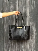 Cartera tipo tote bag de cuero pelo negro con herrajes dorados, doble asa de hombro y detalle de flecos colgantes.