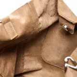 Cartera tipo tote bag de gamuza en color tostado, con doble asa de mano y correa ajustable extraíble. Presenta detalles de tachas metálicas en los laterales y un logo circular grabado en el centro.