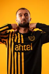 Camiseta de fútbol Puma del Club Atlético Peñarol, modelo Aniversario de manga larga. Diseño combinado con la mitad derecha a rayas verticales amarillas y negras, y la mitad izquierda totalmente negra con detalles en amarillo.