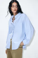 Camisa de manga larga color celeste claro, confeccionada en popelín de algodón. Presenta cuello solapa con botones y un bolsillo de parche en el pecho izquierdo.