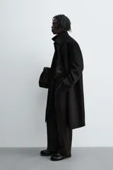 Abrigo negro de corte recto confeccionado en tejido con lana. Tiene cuello alto, manga larga con puños abotonados, bolsillos laterales y abertura en la espalda. Cierre frontal con botones.