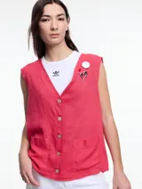 Chaleco sin mangas color coral, con escote en V, cierre frontal con botones de madera y bolsillos de parche. Incluye tres pins decorativos: un corazón plateado, un círculo plateado y un cuadrado rojo. Confeccionado en tejido liviano de lino y viscosa.