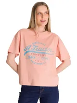 Remera de corte boxy y relajado, color rosado, con cuello redondo y mangas cortas. Presenta un estampado gráfico frontal de estilo retro con tipografía en tonos azules y rojos.