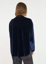 Camisa de terciopelo color bordó, con cuello mao y cierre frontal de botones. Presenta detalles de volados verticales a ambos lados de la botonera y mangas largas con puños abotonados.