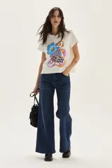 Remera de algodón color blanco, manga corta, con un estampado central que incluye una flor azul y formas abstractas con patrón a cuadros en tonos naranjas y azules, con texto bordado.