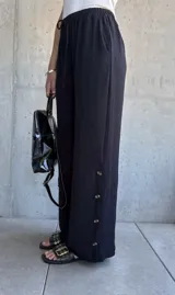 Pantalón negro de corte recto y pierna ancha, con cintura elástica ajustable con cordón y detalle de botones dorados en el lateral inferior.