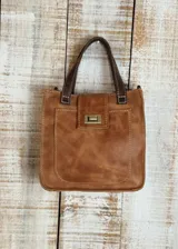 Mini cartera de cuero color tostado con costuras visibles en contraste. Presenta un bolsillo frontal con cierre metálico tipo torniquete, doble asa de mano en cuero oscuro y estructura rígida.