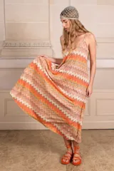 Vestido largo tipo solera con diseño plisado y estampado geométrico en zigzag en tonos naranja, crema y gris. Presenta breteles finos y una silueta holgada y fluida.