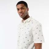 Camisa de manga corta color blanco con diseño estampado de pequeñas palmeras negras, cuello camisero y cierre frontal de botones.
