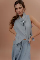 Chaleco de lino color marfil o beige claro, con diseño sin mangas, cuello alto amplio, cierre frontal con botones y bolsillos delanteros amplios. Presenta un corte asimétrico y relajado.