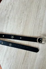 Cinturón de cuero vacuno negro con hebilla metálica plateada ovalada y apliques decorativos en forma de estrella metálica.