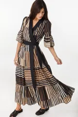 Vestido largo de algodón con patrón de rayas verticales y bloques de colores en tonos marrón, negro y crudo. Presenta diseño cruzado tipo envolvente con solapas negras, mangas tres cuartos acampanadas con volados, falda escalonada y lazo negro ajustable en la cintura.