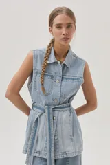 Chaleco largo de jean celeste con cuello camisero, sin mangas, con bolsillos frontales, laterales e internos, y lazo ajustable en la cintura.
