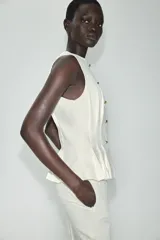 Chaleco entallado blanco con cuello redondo, manga sisa, pinzas estructuradas, detalle de peplum en la parte inferior y cierre frontal con botones dorados.