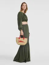 Bolso tipo tote pequeño de paja trenzada color natural, con ribetes y asas de cuero rojo. Presenta un logo bordado de dos caballos galopando en color rojo en el centro.