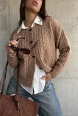 Cardigan de punto trenzado en color beige, con cuello redondo, cierre frontal de botones oscuros y mangas largas con puños acanalados.