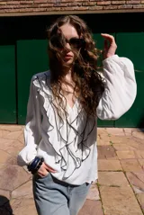 Blusa blanca de manga larga con volados en el frente y bordes en contraste negro. Presenta escote en V y mangas abullonadas con puños elásticos.