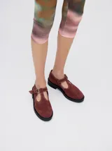 Zapatos tipo guillermina de gamuza color bordó, con diseño de tira en T, cierre con hebilla metálica y costuras a la vista en hilo blanco. Poseen una suela de plataforma de goma negra de 4 cm.