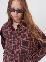 Camisa de seda con cuello camisero, manga larga y bolsillo frontal. Presenta un diseño estampado geométrico de cuadrados en tonos marrones y azul oscuro.