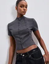 Top de sastrería color gris grafito, con cuello alto, manga corta y silueta entallada. Presenta un diseño con doble hilera de botones decorativos frontales y largo a la cintura.