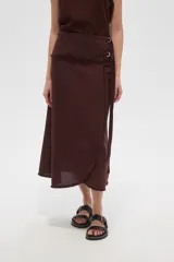Falda larga de algodón color marrón oscuro, de corte envolvente (wrap skirt) con abertura lateral y borde deshilachado. Se ajusta con un lazo lateral que pasa por argollas metálicas.