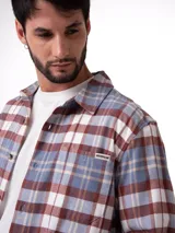 Camisa de manga larga con estampado de cuadros en tonos gris, azul y marrón. Presenta cuello camisero, cierre frontal con botones, bolsillo en el pecho con ranura para bolígrafo y dobladillo redondeado.