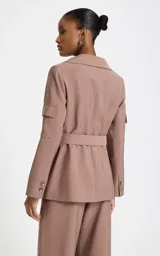 Blazer sastre color marrón claro con patrón de raya diplomática, cuello con solapa ancha, mangas largas con bolsillos aplicados en las mangas, bolsillos frontales con solapa, cinturón incorporado con hebilla metálica y cierre cruzado.