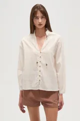 Camisa de algodón color blanco, con cuello en V, cierre frontal con botones y cordones decorativos con mostacillas en el escote. Presenta mangas largas con puños y un diseño de textura sutil.