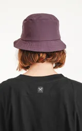 Piluso de estilo bucket hat en color violeta oscuro, con costuras circulares en el ala y parche cuadrado frontal con logo de tijeras.