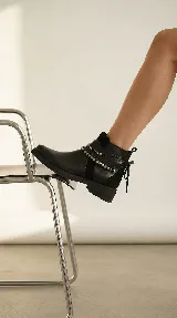 Bota corta de cuero negro con taco cuadrado bajo. Presenta un diseño versátil con un accesorio desmontable que incluye flecos de cuero, tachas metálicas y una cadena decorativa alrededor del tobillo.