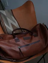 Bolso de viaje tipo weekender de cuero color tostado con asas y detalles en marrón oscuro, herrajes metálicos y bolsillo frontal con cierre.