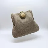 Bolso de yute de algodón en color beige, con herrajes metálicos dorados opacos y un detalle circular con piedra en el centro. Cuenta con cierre de broche de presión y asa de cadena desmontable para usar al hombro.