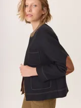 Chaqueta de paño color azul marino con textura de espiga, corte clásico y cuello redondo. Presenta pespuntes contrastantes en color camel, bolsillos frontales de parche y coderas de gamuza color camel. Cierre frontal con cremallera dorada.