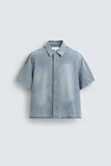 Camisa de corte boxy fit confeccionada en denim ligero de algodón. Presenta cuello solapa, manga corta y cierre frontal con botones. Diseño con efecto lavado.