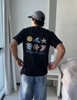 Remera negra de manga corta con estampado gráfico en la espalda que incluye un sol amarillo, una estrella de mar, una flor, una ola con el logo 'Brokers' y formas abstractas en tonos pastel, junto al texto 'VITAMIN SEA'.
