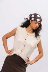 Chaleco de punto color beige, sin mangas, con cuello redondo y cierre frontal con botones de madera. Presenta un tejido de punto liso y terminaciones acanaladas en cuello y sisas.