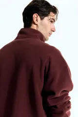 Buzo deportivo de polar color bordó, con cuello alto y cierre frontal parcial. Presenta mangas largas de corte caído y bolsillos laterales integrados en la costura.