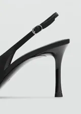 Zapato de tacón destalonado color negro de tejido satinado, con punta fina, cierre de hebilla ajustable en la tira del talón, tacón de aguja y detalle de flor decorativa en el empeine.