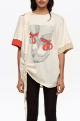 Remera larga de color marfil con detalle de abertura y lazos en el hombro, estampa frontal de zapatillas de ballet con lazos rojos y mangas cortas con puños en contraste rojo y beige. Cuenta con cordón ajustable lateral.