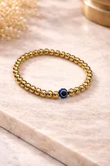 Pulsera elástica compuesta por cuentas esféricas doradas y un dije central con forma de ojo turco (azul y blanco).