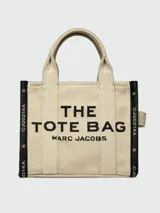 Bolso tipo tote Marc Jacobs modelo The Small Tote, color beige con detalles y texto en negro que dice "THE TOTE BAG MARC JACOBS". Cuenta con asas superiores y laterales para llevar en la mano o al hombro.