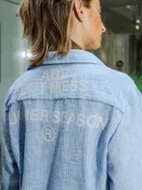 Camisa de lino rayada en tonos celeste y blanco, con cuello y botones frontales. Presenta un estampado en el frente y la espalda.