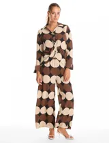 Conjunto de dos piezas compuesto por camisa de manga larga con nudo frontal y pantalón estilo palazzo de pierna ancha. Ambas prendas presentan un estampado geométrico de círculos grandes en tonos marrón, negro y beige. La camisa cuenta con cuello solapa y cierre frontal con botones, mientras que el pantalón posee tiro alto y corte holgado.