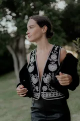 Chaleco negro con escote en V profundo, confeccionado en tela con bordados florales en hilo plateado. Presenta un diseño estructurado con ribetes plateados en los bordes y cierre frontal.